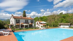 A swimming pool with a tiled deck, surrounded by two-story houses with red-tiled roofs, and a lush green hillside in the background. - Podere Ritali by Interhome (Civitella in val di Chiana)