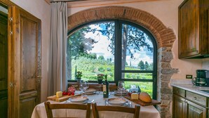 A dining area with a table set for two, a view of a vineyard through a large arched window, and wooden cabinetry. - San Rocco by Interhome (Greve in Chianti)