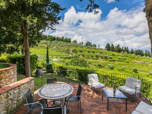 A patio with a round table and chairs, surrounded by a stone wall and a view of a green hillside. - San Rocco by Interhome (Greve in Chianti)