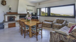 Ein geräumiges Wohnzimmer mit Kamin, einem Esstisch mit Stühlen, einer Sofagarnitur und einem großen Fenster mit Blick auf die Landschaft.
