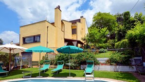 Ein Schwimmbecken mit Liegestühlen und Sonnenschirmen, umgeben von einer gepflegten Rasenfläche und einem Gebäude mit gelber Fassade.