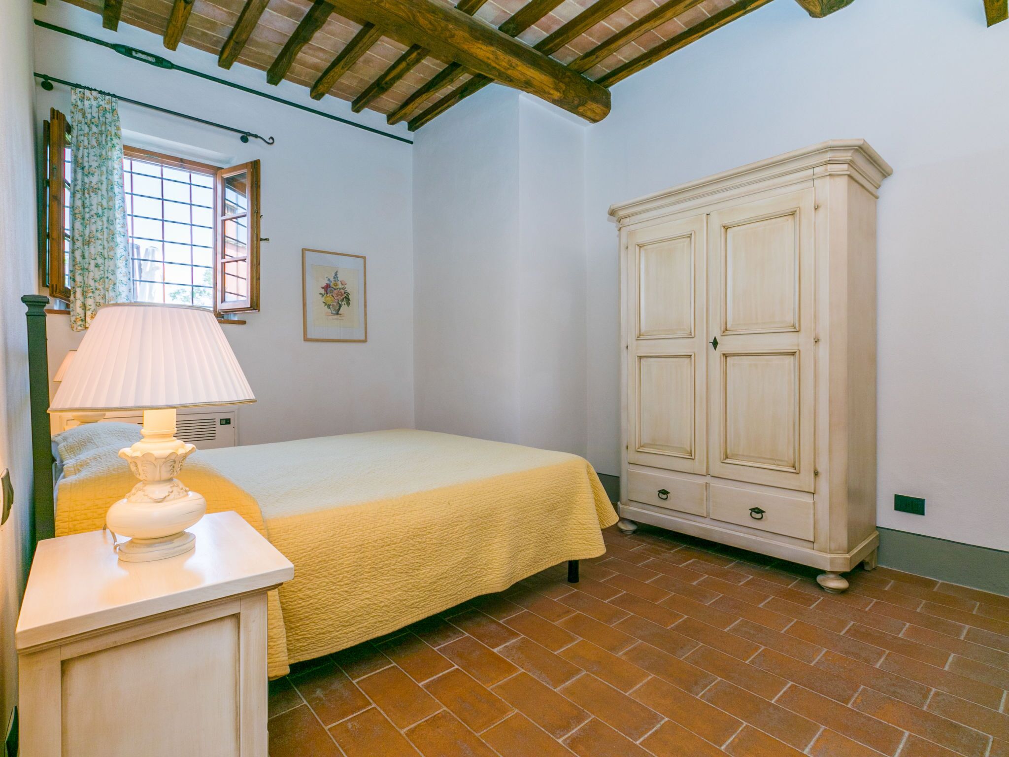 Un dormitorio con cama, una mesita de noche con lámpara, un armario, una ventana con cortinas y un cuadro en la pared.