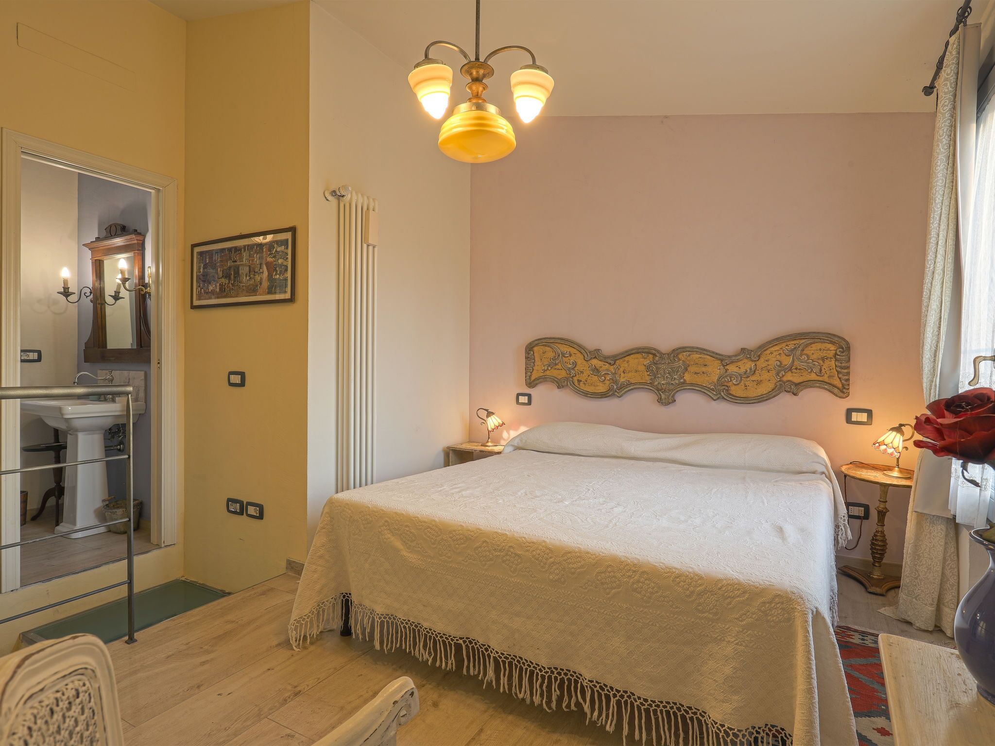 A hotel room with a bed, a bedside table with a lamp, a mirror, a radiator, and a picture on the wall.