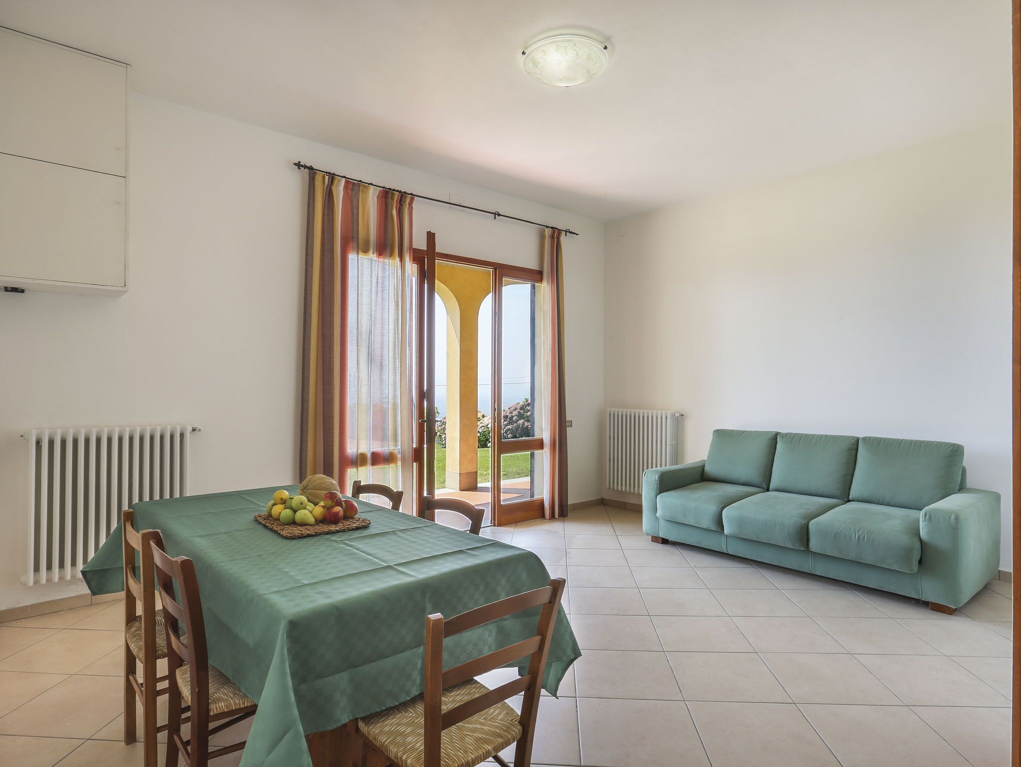 Un coin repas avec une table dressée pour deux personnes, une nappe verte et un panier de fruits. On y trouve également un canapé vert et une fenêtre avec des rideaux.
