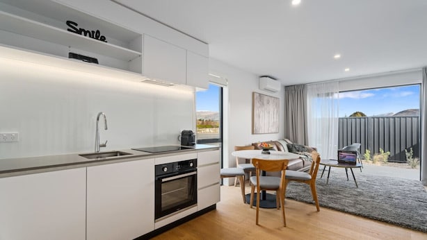 Una cocina moderna con armarios blancos, horno empotrado y un lavamanos. Hay una zona de comedor con una mesa redonda y sillas, y un amplio ventanal con vistas al exterior.