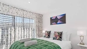 A modern bedroom with a green upholstered bed, a large window with sheer curtains, and a painting on the wall. - Explore Wanaka - Mountain Views, Riverside Locale (Albert Town)