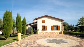 A house with a stone-paved entrance, a flower design on the ground, and a well-maintained garden with tall trees.