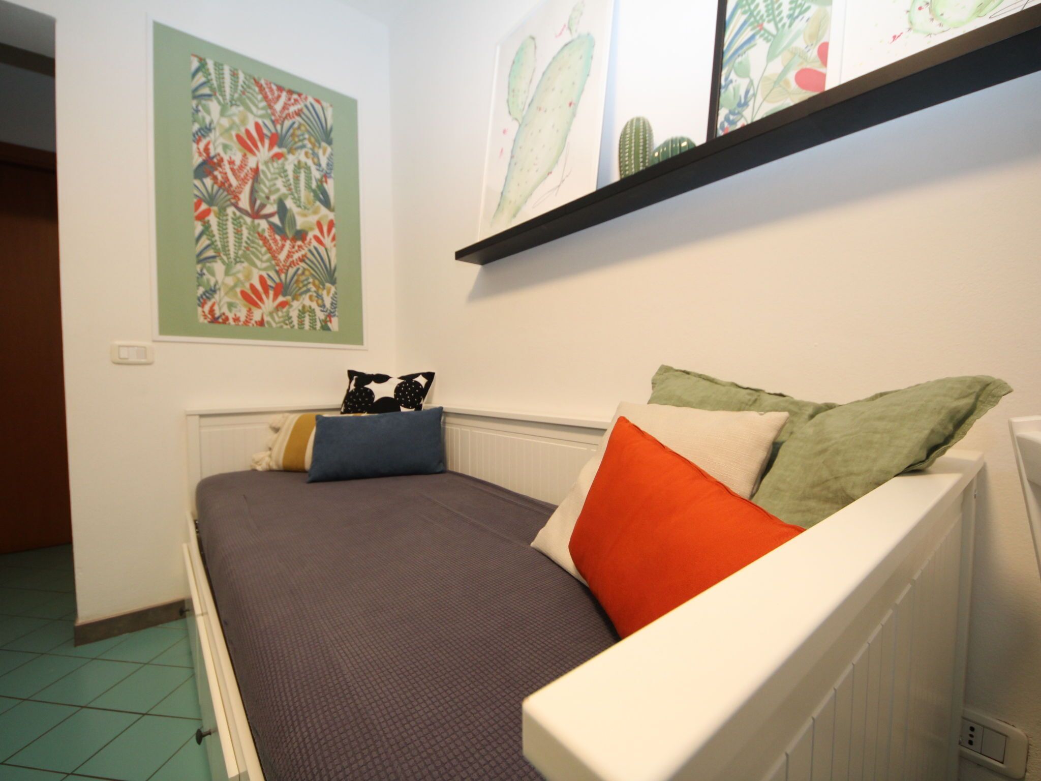 Een kleine, verzorgde slaapkamer met een enkel bed, een plank boven het bed en een kleurrijk schilderij aan de muur.