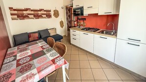 A compact kitchen with white cabinets, a gas stove, and a sink. A dining table with a patterned tablecloth is set against the wall. - Solmare by Interhome (Rosolina Mare)
