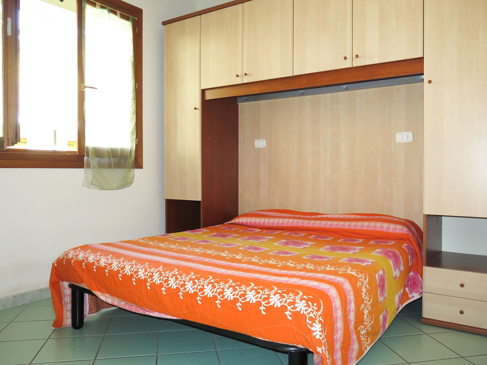 Een slaapkamer met een bed, een kledingkast, een raam met gordijnen en een betegelde vloer.