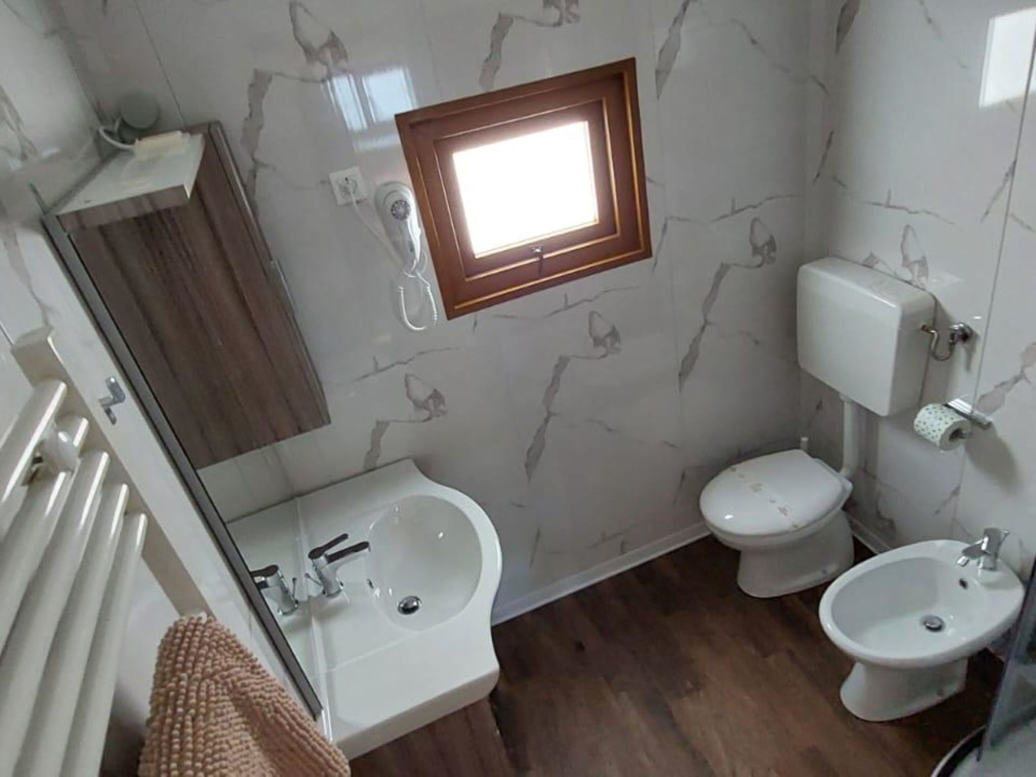 A bathroom with a marble wall, a wooden-framed window, a toilet, a sink, and a bidet.