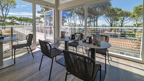 Una terraza soleada con mobiliario exterior, ventanas amplias y vistas a árboles y un cuerpo de agua.