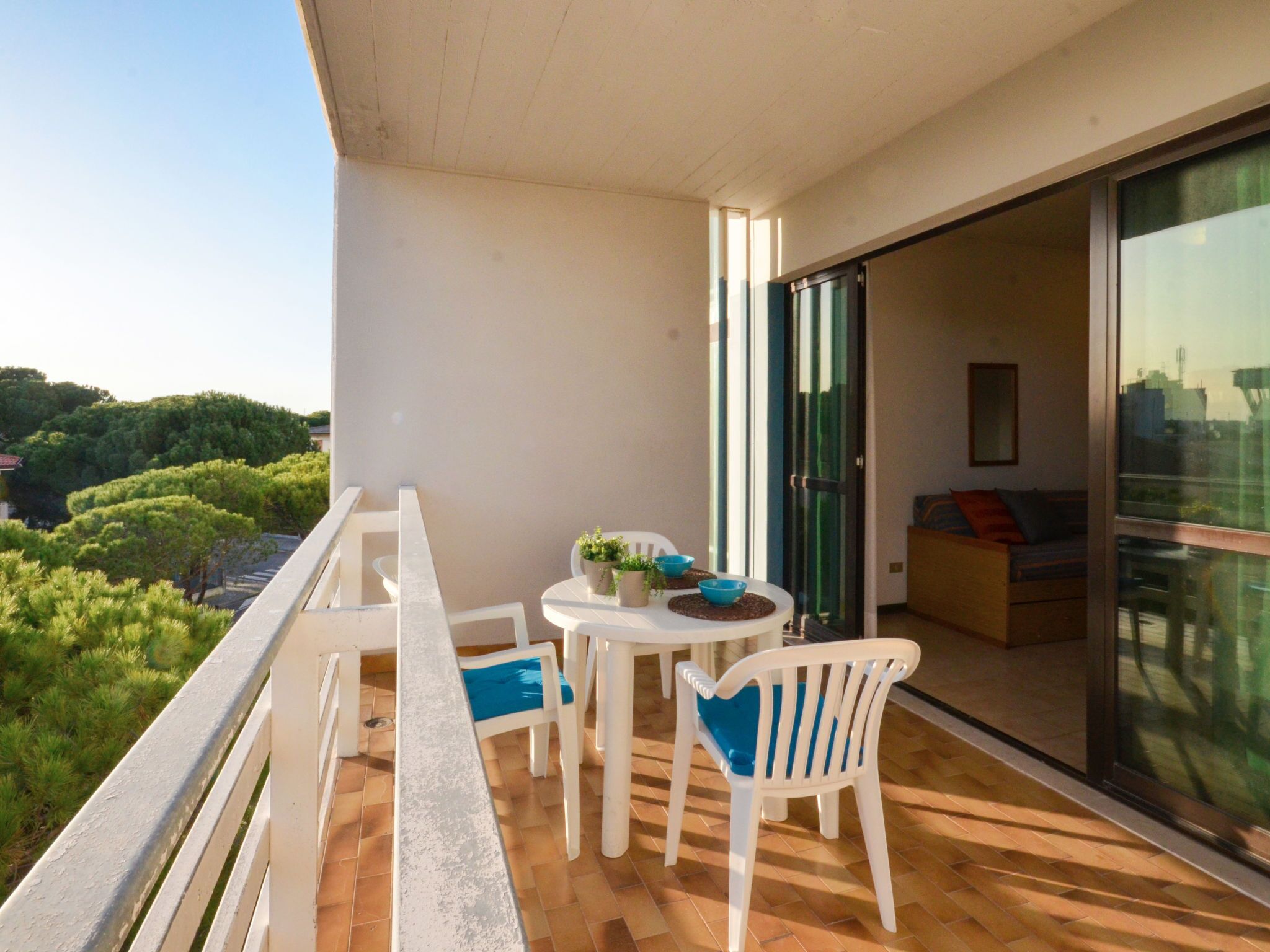 Ein Balkon mit weißem Tisch und Stühlen, einem kleinen Topfpflanzen und Blick auf Grünflächen und Gebäude.