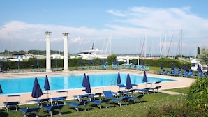 Una piscina con sillas y sombrillas azules, rodeada de vegetación y con vista a barcos y yates al fondo.
