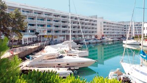 Un puerto deportivo con varias embarcaciones amarradas, un edificio de varios pisos con balcones, y un cielo despejado.