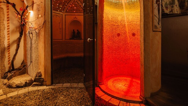 Una habitación con paredes de piedra, un área circular iluminada con luz roja, una puerta de madera y un banco de piedra.