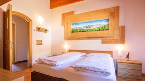 Un dormitorio con dos camas, cabecero de madera, un cuadro de montañas, lámparas de noche y una mesita de noche con un adorno.