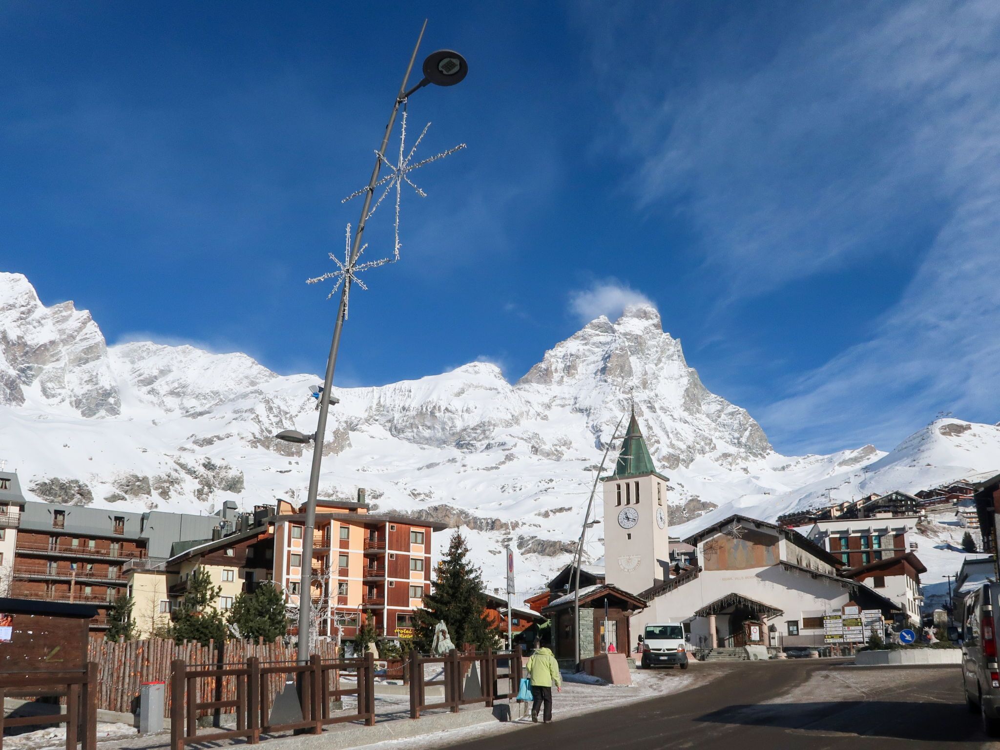Un pueblo nevado a los pies de una montaña, con una imponente torre de iglesia, edificios con balcones y un cielo azul despejado.