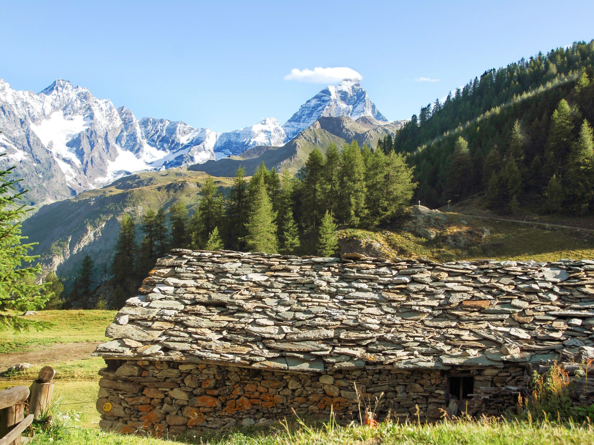 Un bâtiment en pierre surmonté d’un toit en pierre, entouré d’un paysage montagneux avec des sommets enneigés et de vastes forêts.