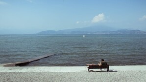 A person sitting on a bench by a calm body of water with a distant shoreline and mountains in the background. - Camping Wien by Interhome (Peschiera del Garda)