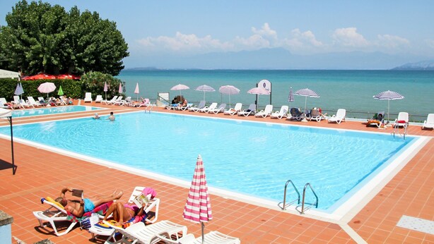 A swimming pool with lounge chairs and umbrellas, surrounded by a tiled deck and a view of the sea. - Camping Wien by Interhome (Peschiera del Garda)