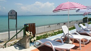 A seaside view with a sign indicating a pedestrian path to the lake, white lounge chairs, and striped beach umbrellas.
