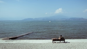 A person sitting on a bench by a calm body of water with a distant shoreline and mountains in the background.