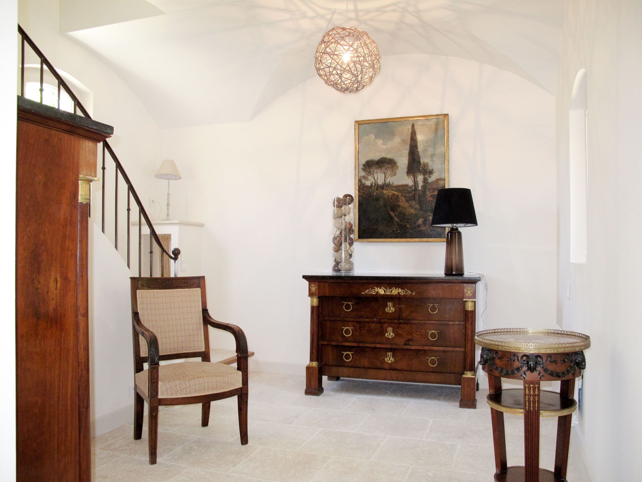 A staircase with a wooden handrail, a chair with a woven seat, a wooden dresser with drawers, a painting on the wall, and a small round table with a lamp.