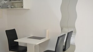 Ett litet rum med ett vitt bord och två svarta stolar. I hörnet står en säng med vita sängkläder.