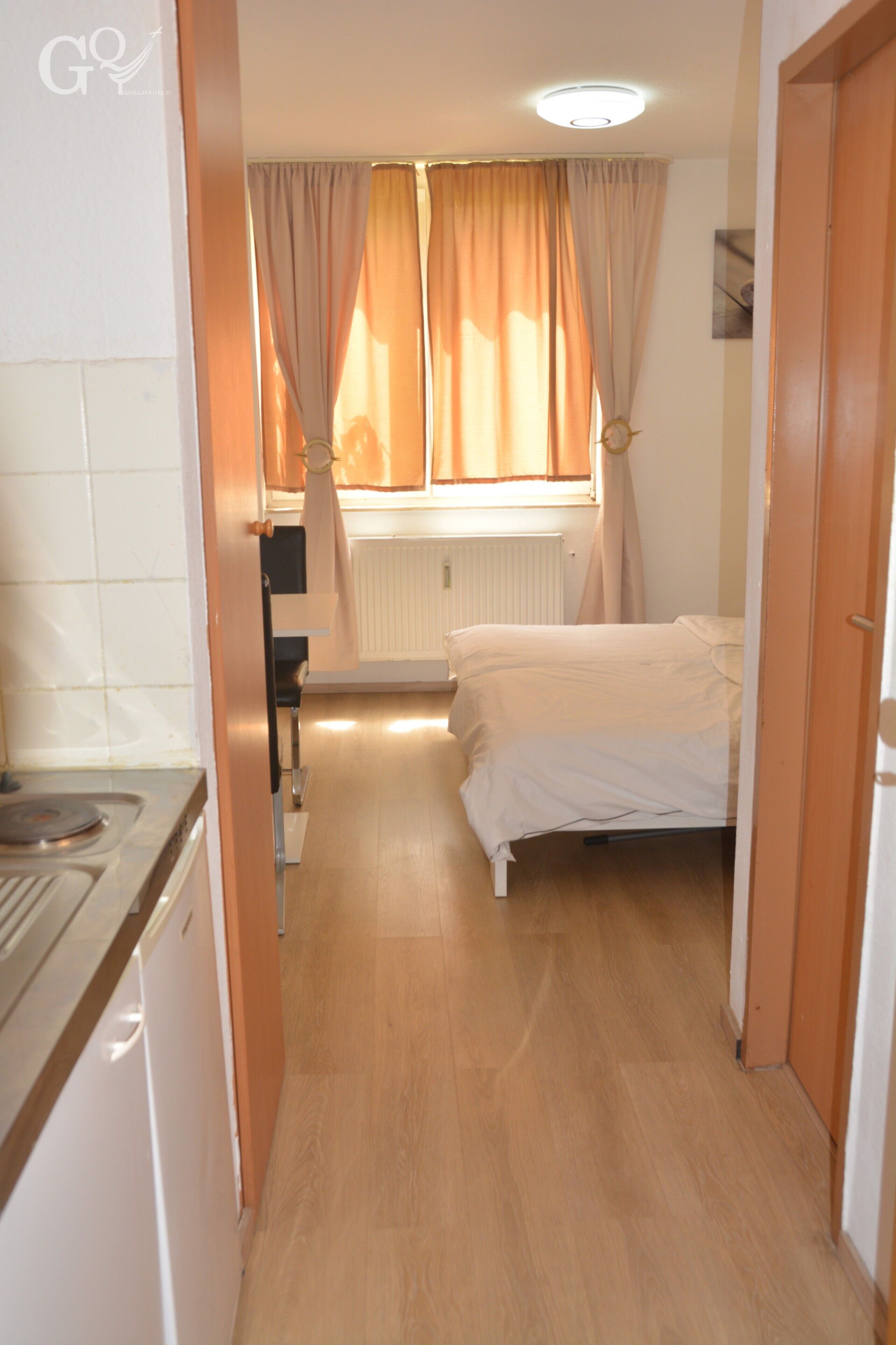 Un couloir étroit mène à une chambre à coucher qui contient un lit, un radiateur et une fenêtre avec des rideaux.