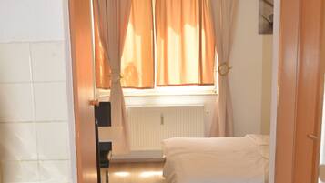 Un couloir étroit mène à une chambre à coucher qui contient un lit, un radiateur et une fenêtre avec des rideaux.