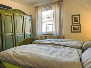 Ein Zimmer mit zwei Betten, einem fensterbehangenen Fenster, einem Heizkörper und zwei gerahmten Bildern an der Wand.