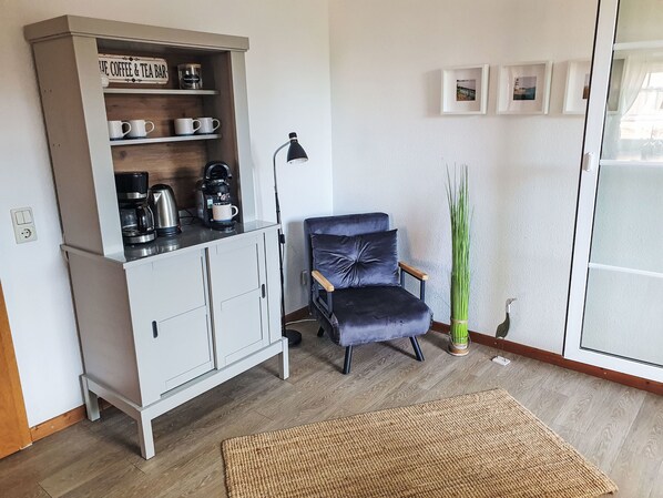 A living room with a coffee and tea bar, a blue armchair, a tall green plant, and a woven rug.