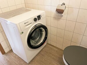 Eine frontgeladene Bauknecht Waschmaschine mit Bedienfeld, ein wandmontierter Papierhandtuchspender und eine Toilette in einem Badezimmer mit gefliesten Wänden und Holzboden.