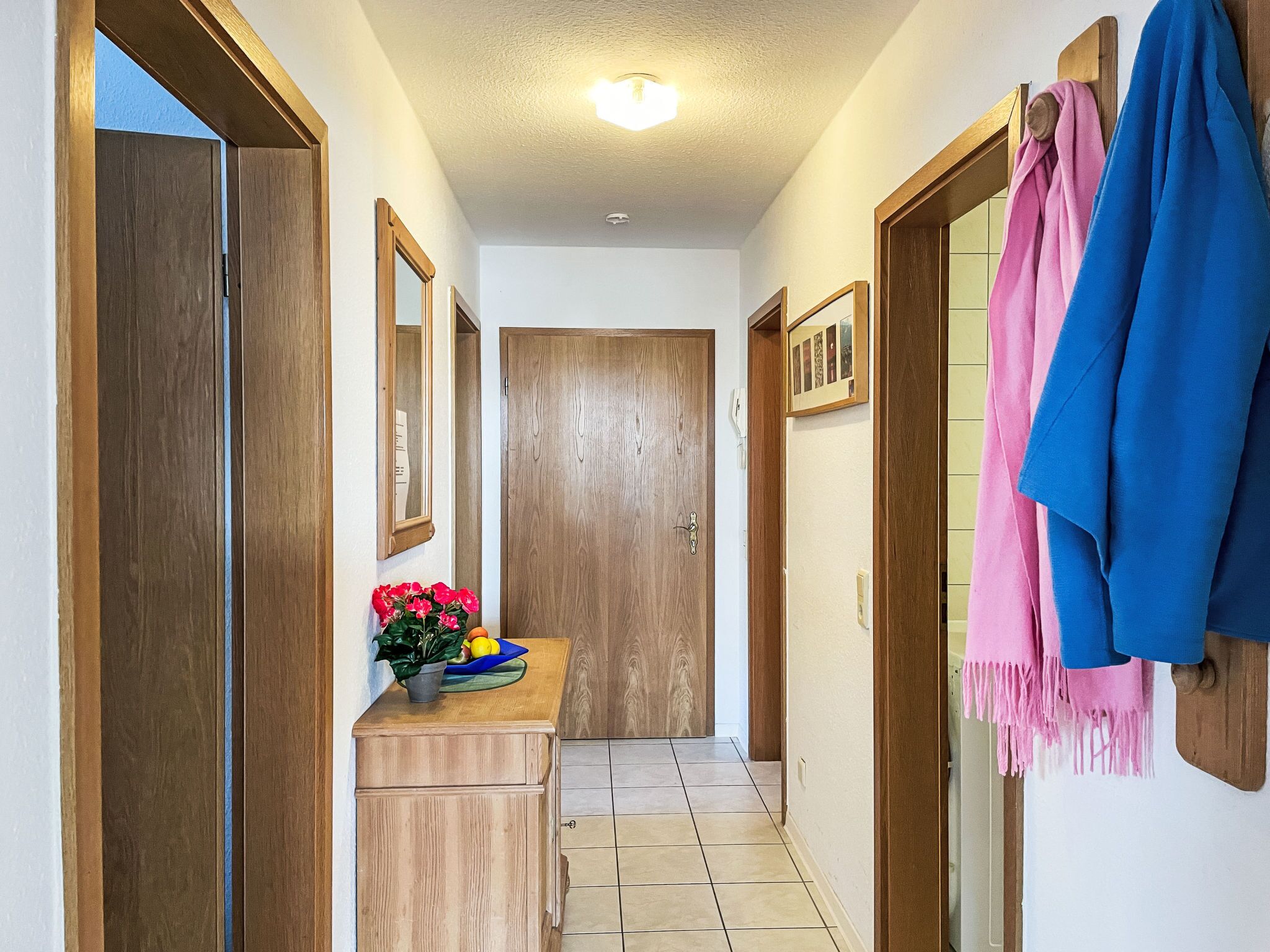 Ein Flur mit Holztüren, einem kleinen Holzschrank mit Blumen in einer Vase und einem Kleiderständer mit einem blauen und rosa Kleidungsstück.