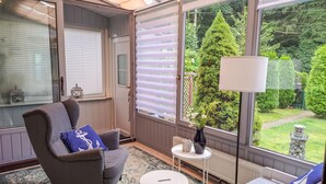 A cozy room with a grey armchair, a small round table, and a sofa. A large window offers a view of a lush garden. - Vacation home Karli by Interhome (Loissin)