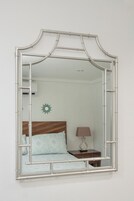 Een spiegel met een bamboe lijst toont een bed, een nachtkastje en een wandlamp.
