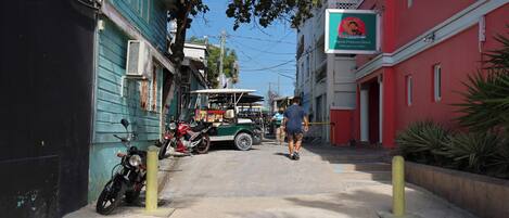 Un carrer estret amb edificis a ambdós costats, una moto aparcada i un tuk-tuk a la distància.