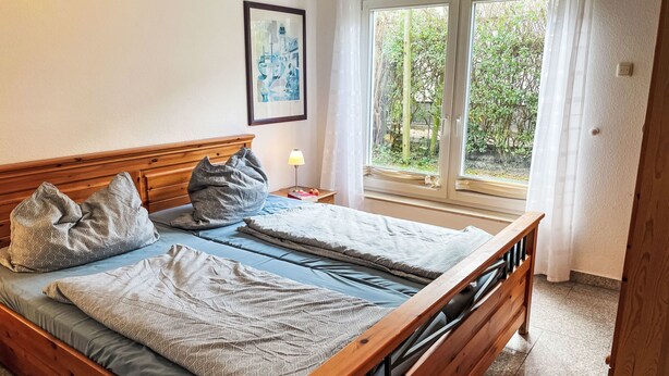 A double bed with patterned bedding, a bedside table with a lamp, a framed picture on the wall, and a window with a view of greenery.
