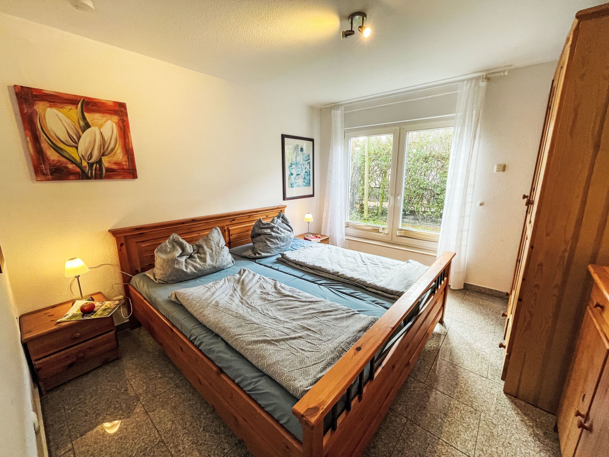 A bedroom with a wooden bed, a nightstand with a lamp, a window with a view of greenery, and a painting on the wall.