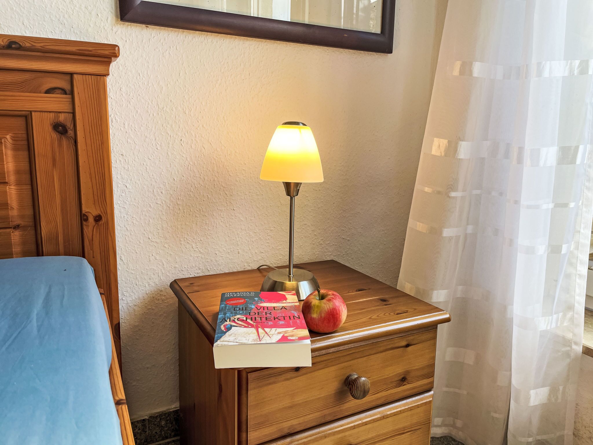 A wooden nightstand with a book titled "Die Villa der Architektur" and an apple, a lamp with a yellow shade, and a blue bedspread.