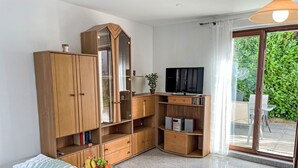 A living room with a glass coffee table, a sofa, a wooden entertainment unit with a TV, and a sliding glass door leading to an outdoor area.