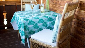 Een houten chalet van binnen, met een eettafel gedekt voor twee personen, een rieten stoel en een vaas met bloemen.