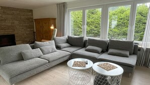 En moderne stue med en grå hjørnesofa, to runde borde med geometriske metalstel, et træskab og store vinduer med udsigt til grøn bevoksning.