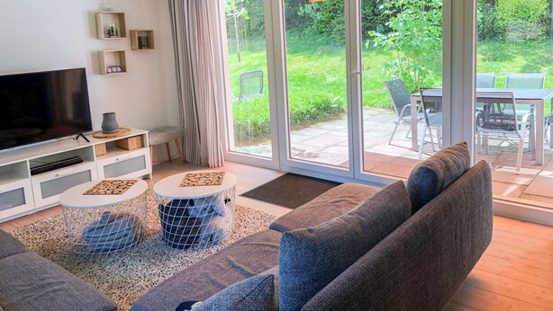 En moderne stue med et fladskærms-tv, en sofa, et sofabord og en skydedør, der fører ud til en udendørs terrasse med et bord og stole.