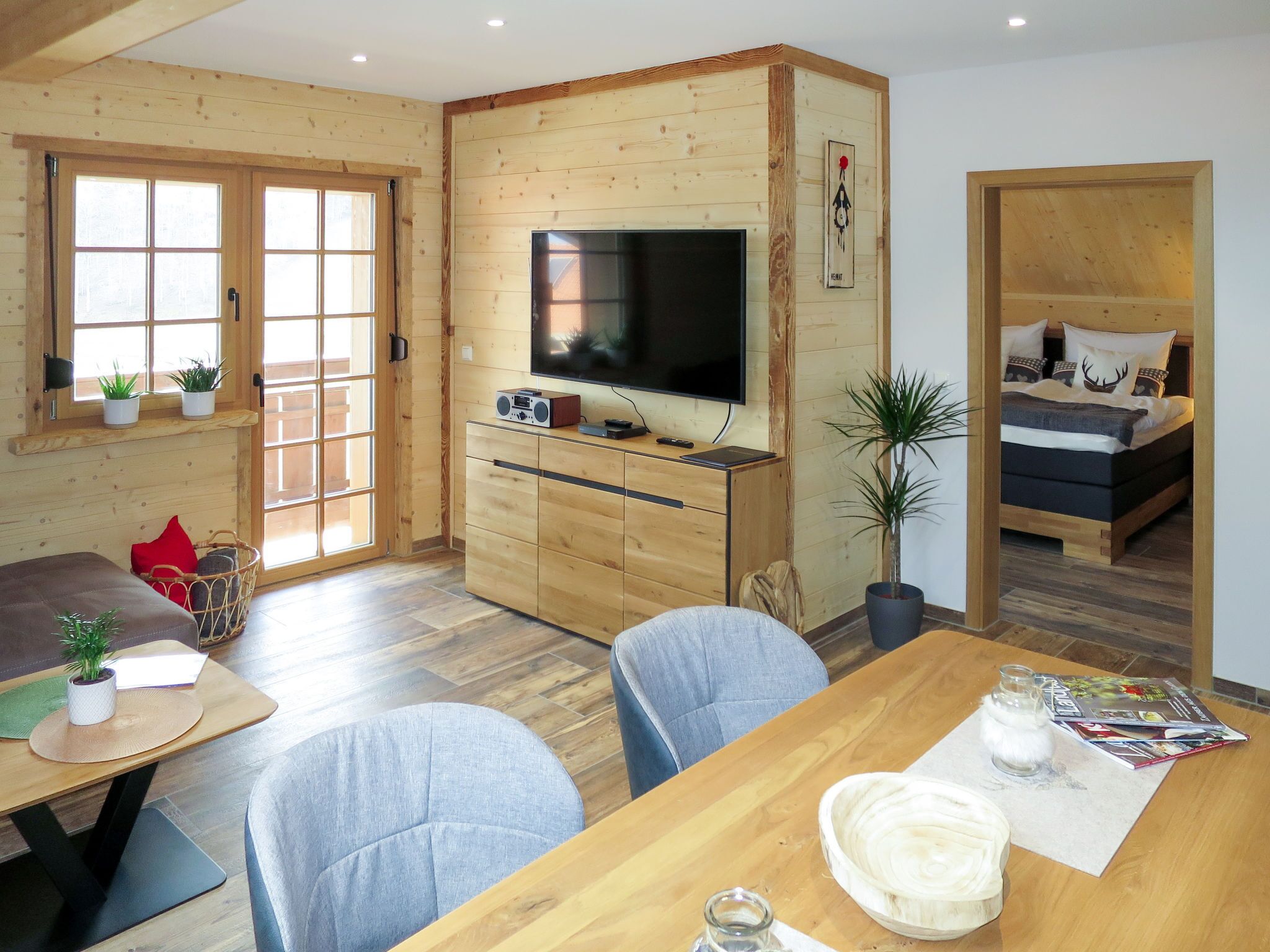 Ein gemütliches Blockhausszimmer mit Holzvertäfelung, einem Esstisch mit Stühlen, einem Fernseher, einem Bett mit Dekokissen und einem Fenster mit Aussicht.