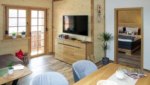 Acogedora habitación tipo cabaña con paredes de madera, una mesa de comedor con sillas, un televisor, una cama con almohadones decorativos y una ventana con vistas.