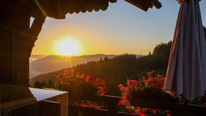 Une vue sur le coucher de soleil depuis un balcon, avec une table et des chaises en bois, donnant sur un paysage montagneux parsemé de fleurs rouges.