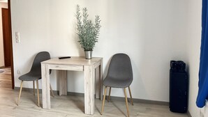 Ett litet, modernt rum med ett träbord, två grå stolar, en krukväxt, en fjärrkontroll och en svart resväska.