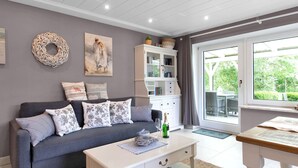 A living room with a grey sofa, white coffee table, and a bookshelf. There is a window with a view of greenery outside. - Deichhaus Küstenwind by Interhome (Fedderwardersiel)
